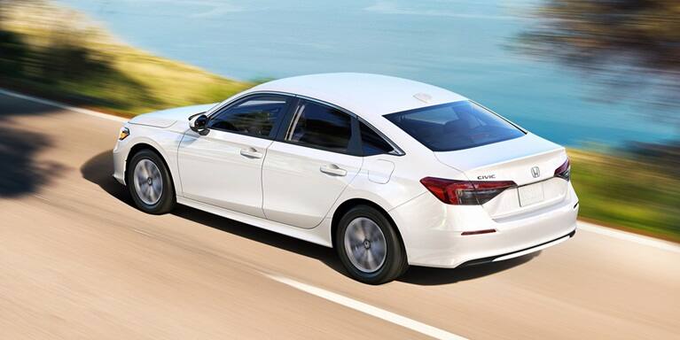 2027 Honda Civic Sedan LX in Platinum White Pearl rear three-quarter view driving on a coastal road (Compact Sedan)