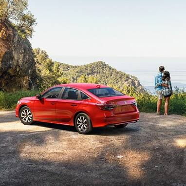 2027 Honda Civic Sedan LX in Rallye Red rear profile parked at a coastal overlook with a couple (Compact Sedan)